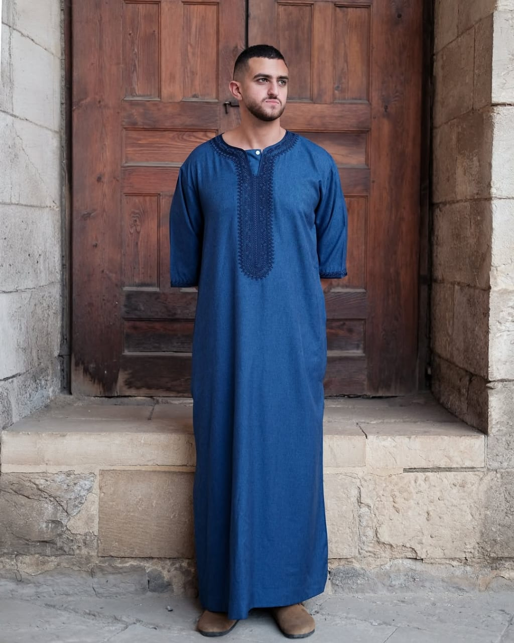 Stabraq blue embroidered thawb photographed in front of a wooden doorway
